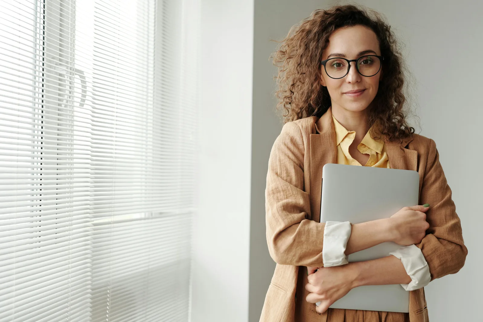 Woman in glasses and a tan blazer holding a laptop by a window
