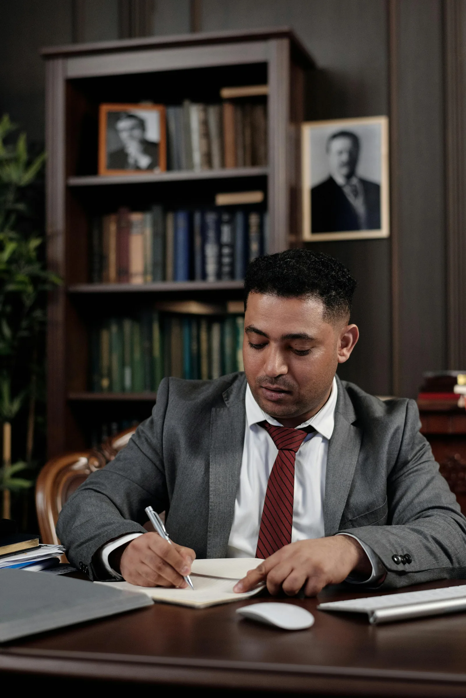 Man in a gray suit writing notes at a desk in front of a bookcase