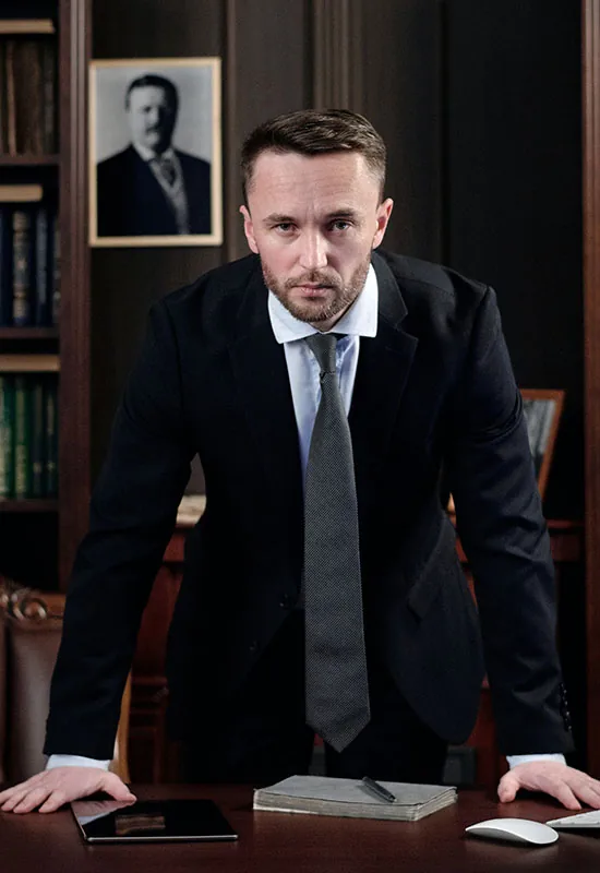 Man in a dark suit leaning on a desk in a wood-paneled office